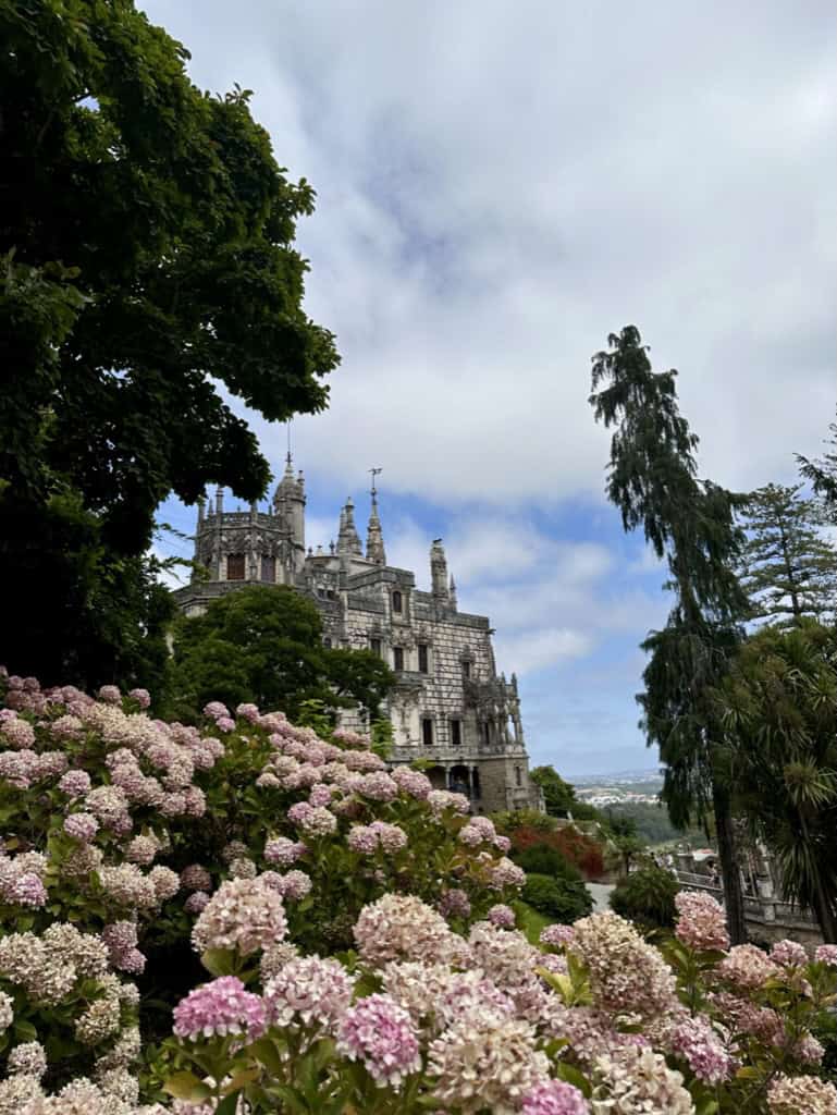 View of the Quinta de Regaleira castle in Sintra, Portugal with green trees and pink flowers.