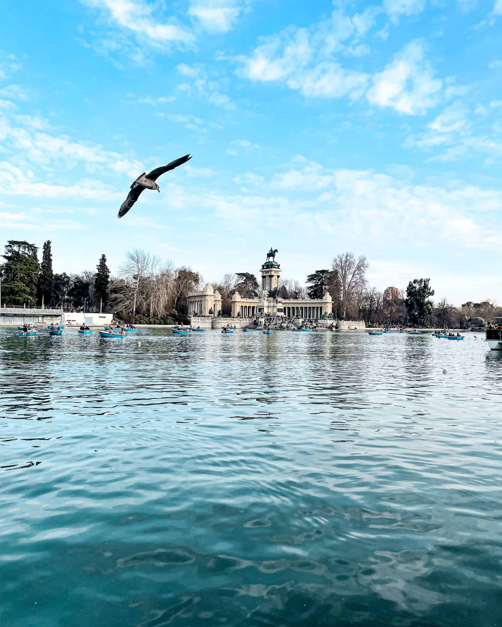 Famous statue and small plaza in The Retiro Park in Spain with a closeup view of the water and people rowing boats and a bird in the sky