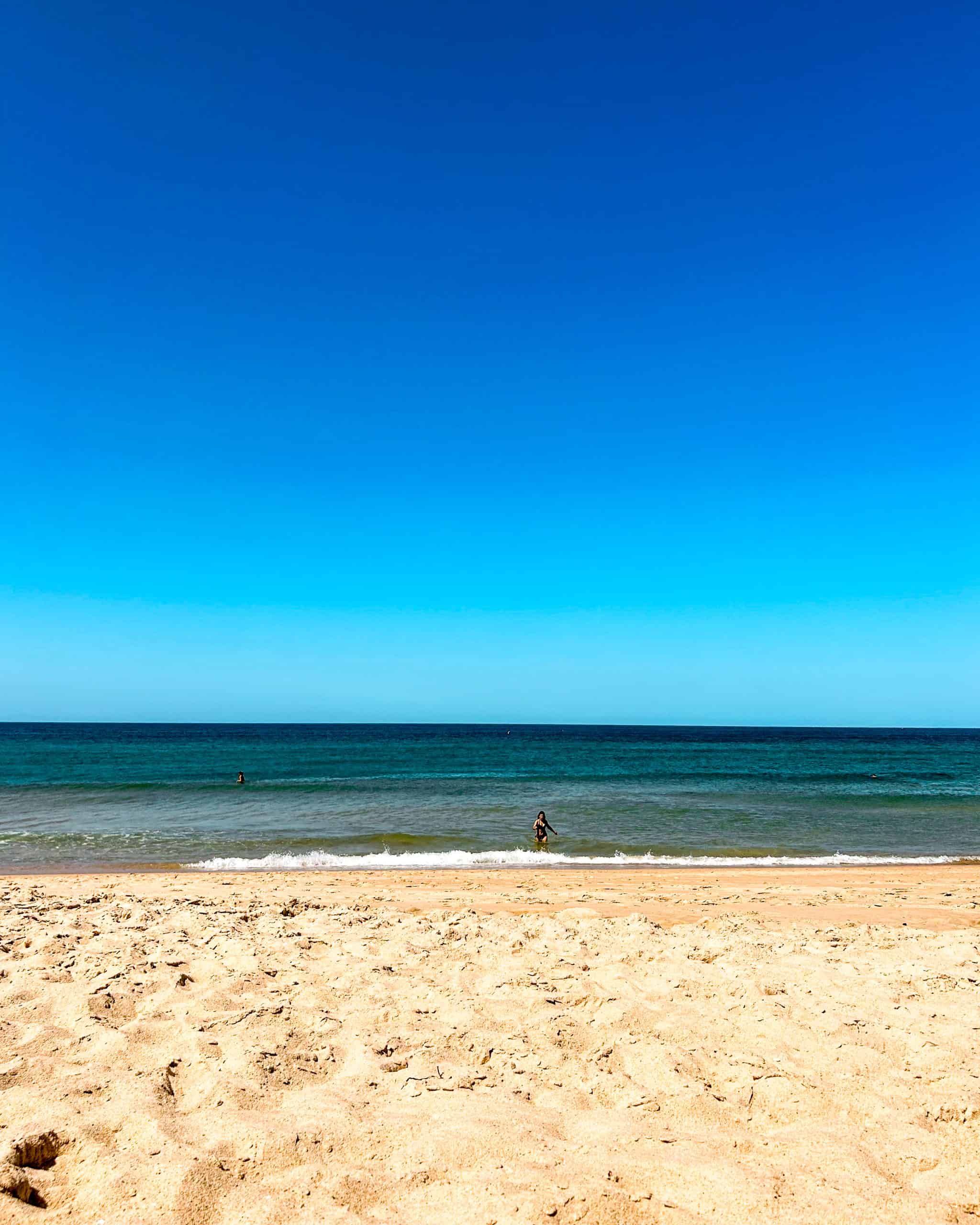 quiet beach with an ocean with no waves featuring one person and a blue sky in Faro, Portugal