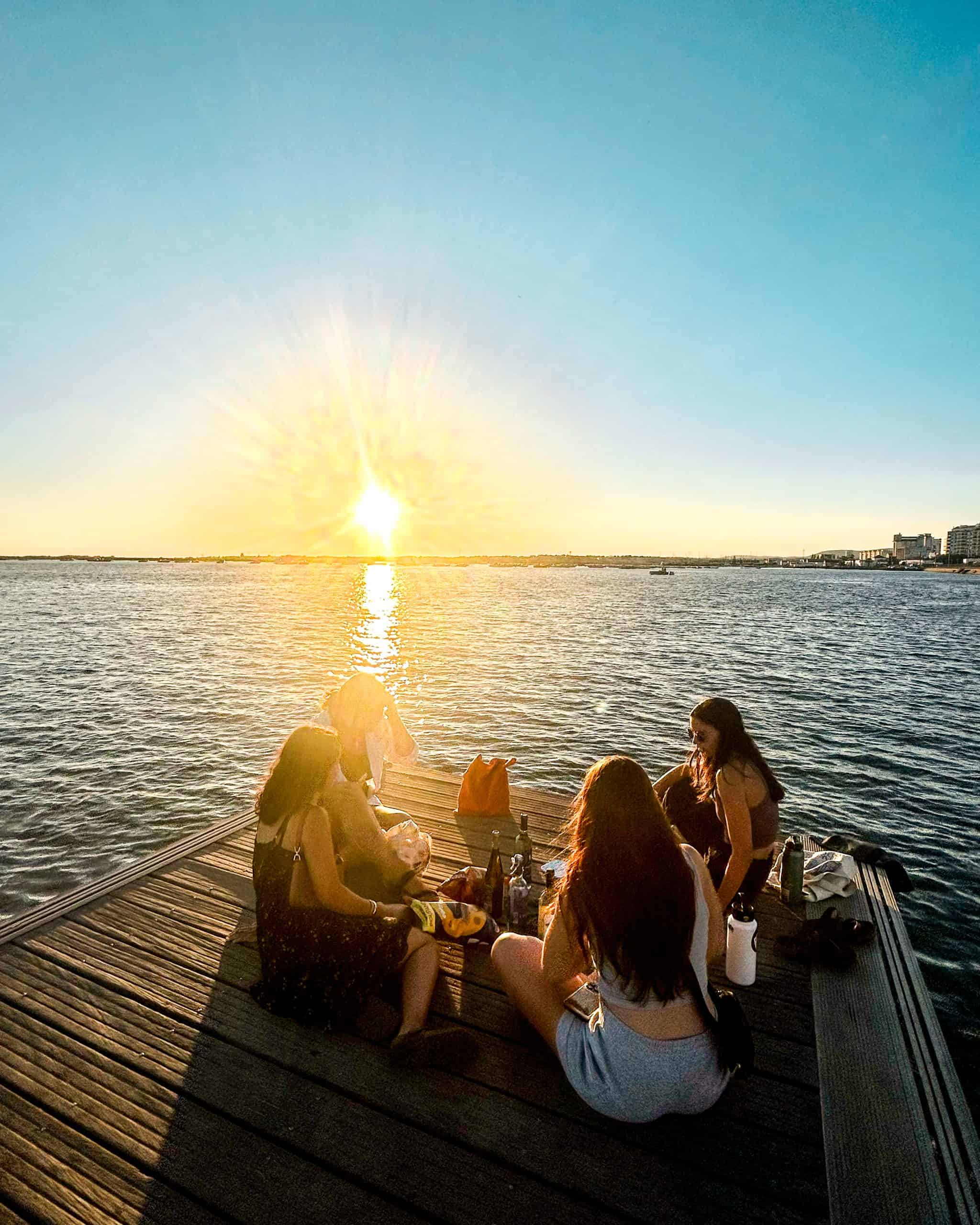 a few women sitting on the docks near the water in Faro, Portugal at sunset