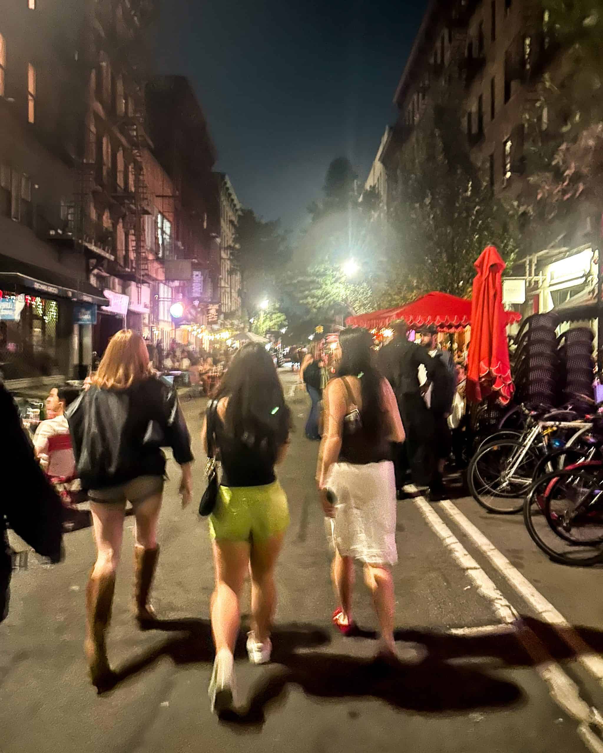 blurry image of three young women walking down the street in the East Village in NYC at night