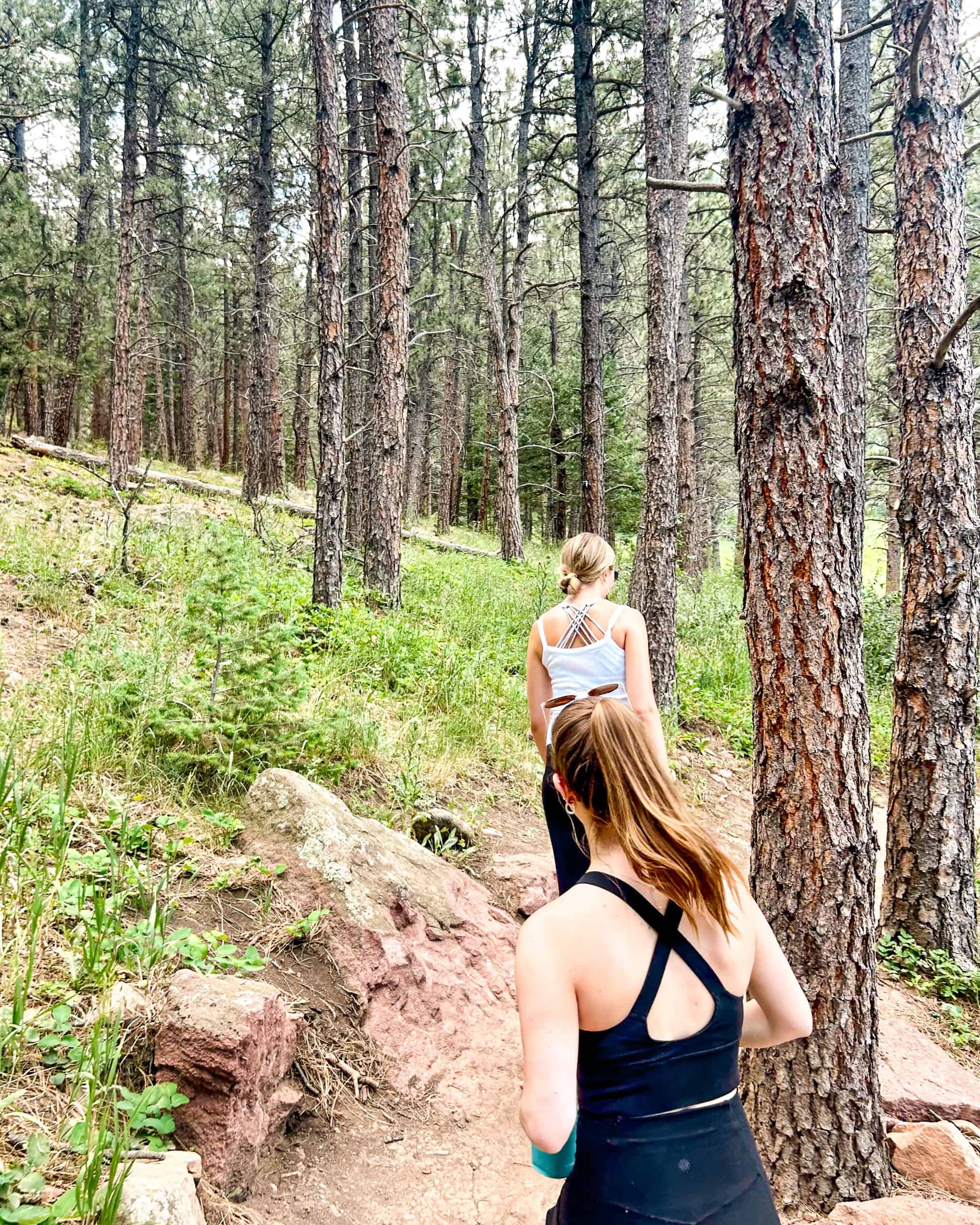 Two women hiking through a forest trail in Boulder, Colorado, surrounded by tall pine trees and rocky terrain