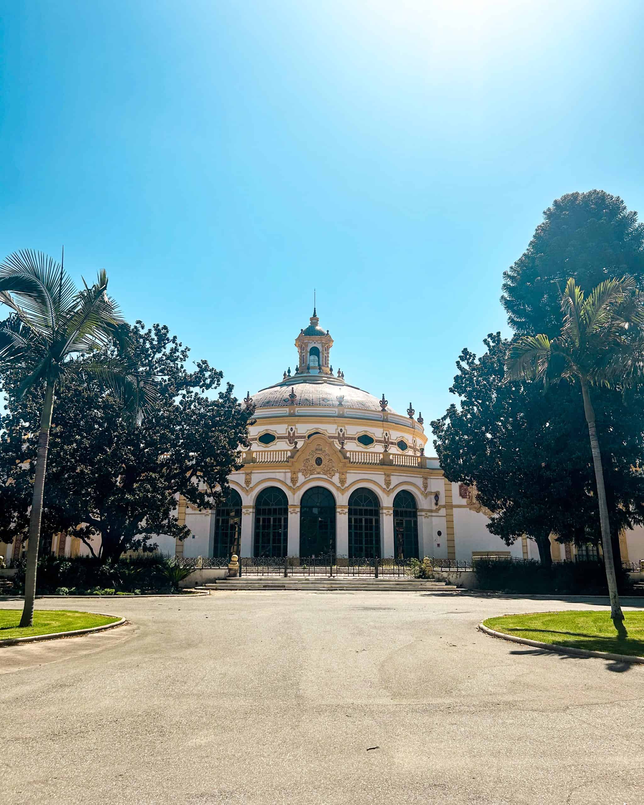 Historic pavilion building inside María Luisa Park in Seville, Spain, with ornate architecture, palm trees, and a wide open courtyard on a sunny day.