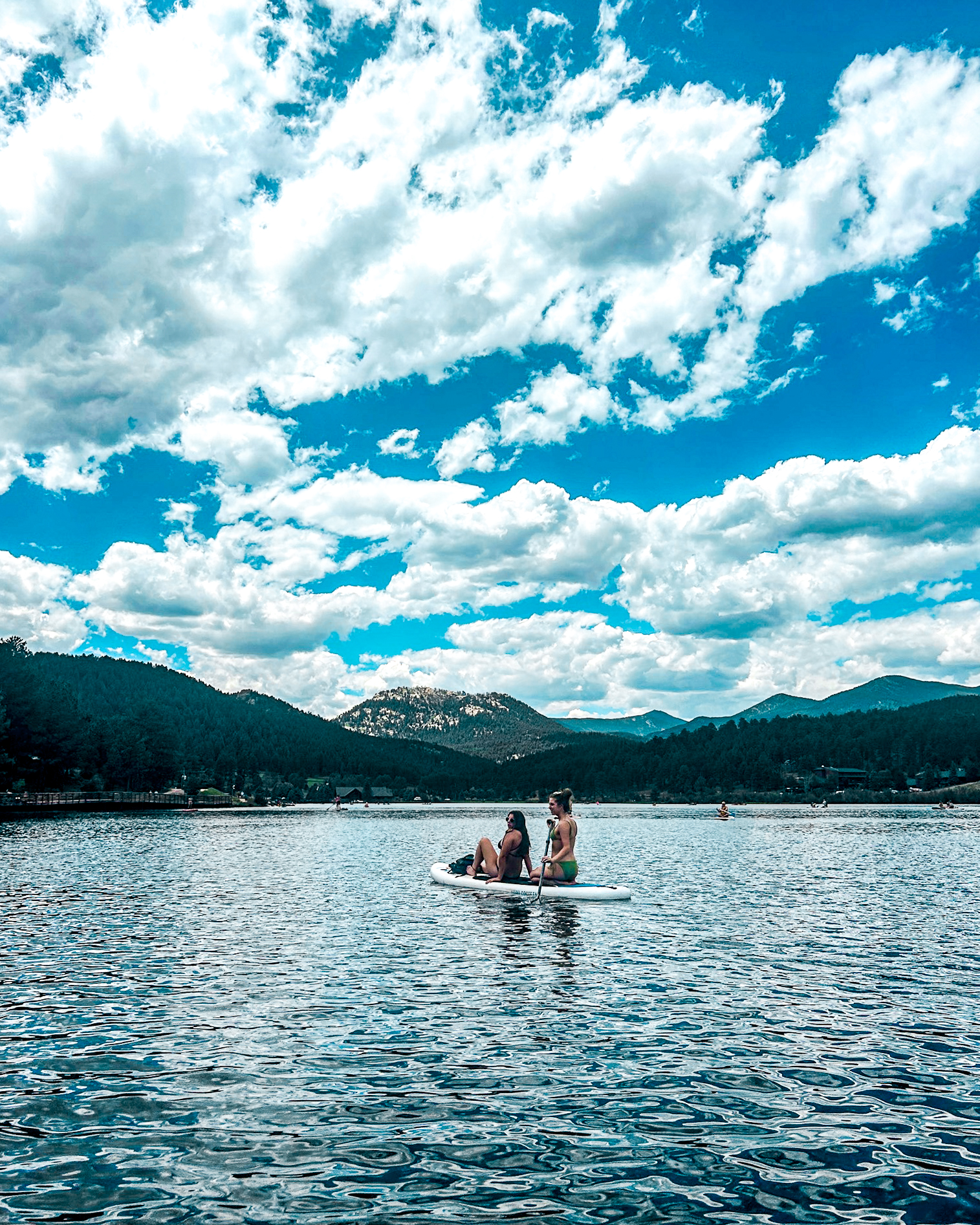 Two women paddleboarding on a calm mountain lake in Colorado, with forested hills, blue sky, and reflections on the water