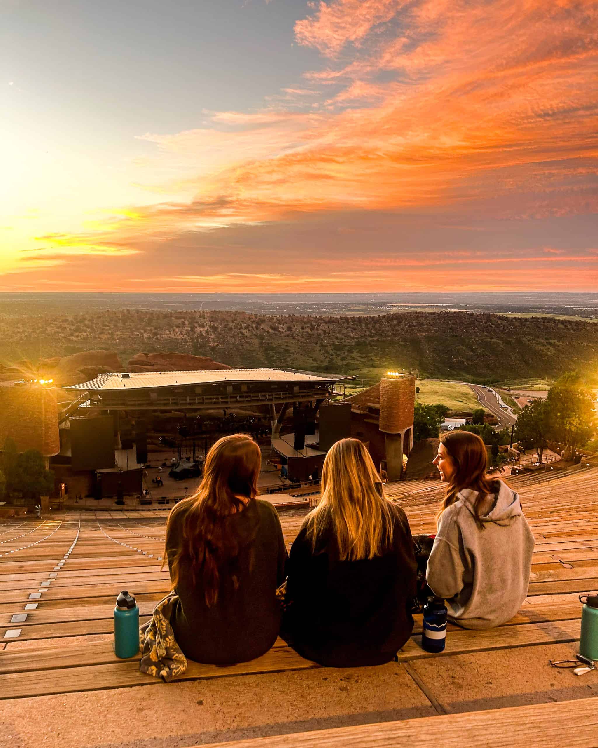 Sunrise view at Red Rocks Amphitheatre in Colorado, with friends sitting on the steps overlooking the stage and dramatic orange sky