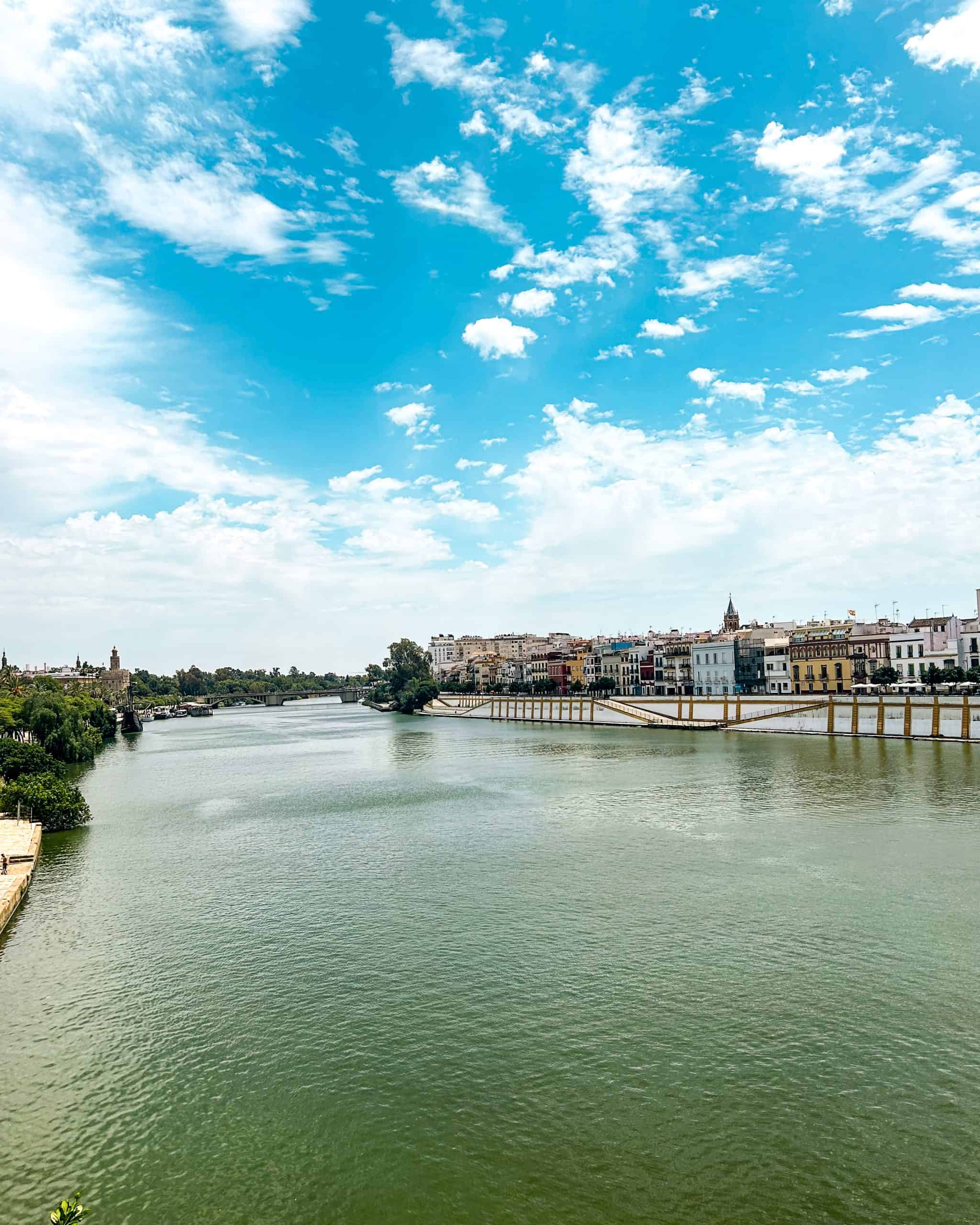 Scenic view of the Guadalquivir River in Seville, Spain, with historic buildings lined along the riverbank and a bright blue sky overhead
