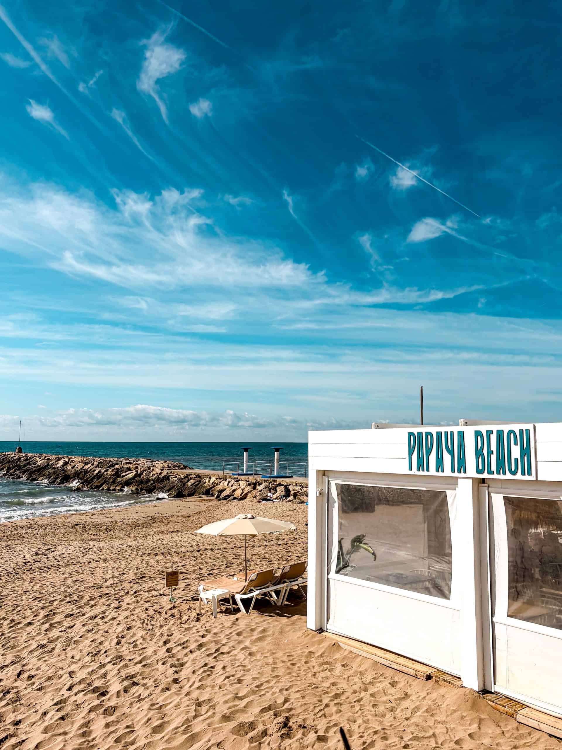 Sunny sand, loungers, and a stone jetty beside the Papaya Beach hut in Sitges
