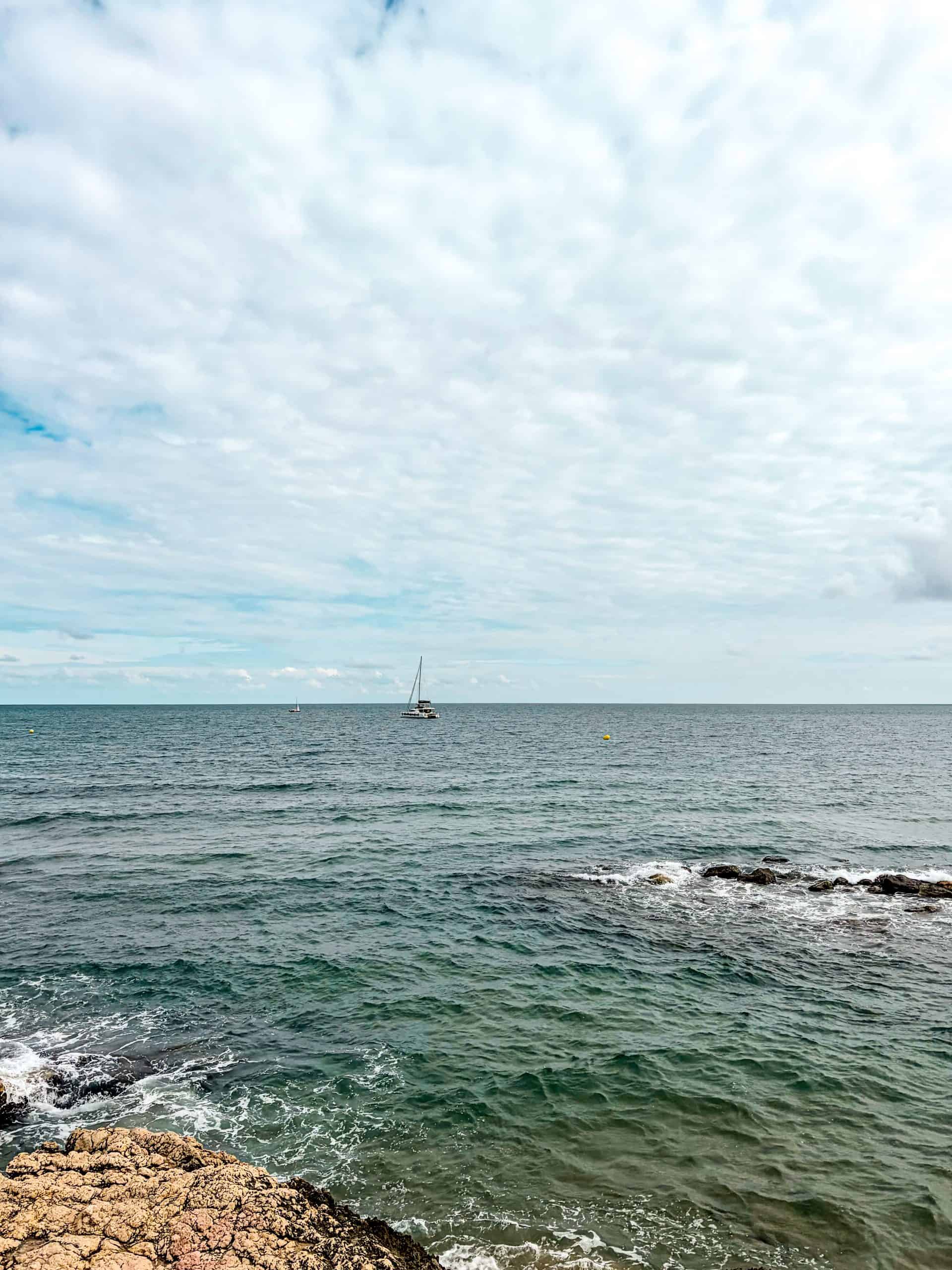 Calm Mediterranean sea off the rocky Sitges shoreline with a catamaran under clouds